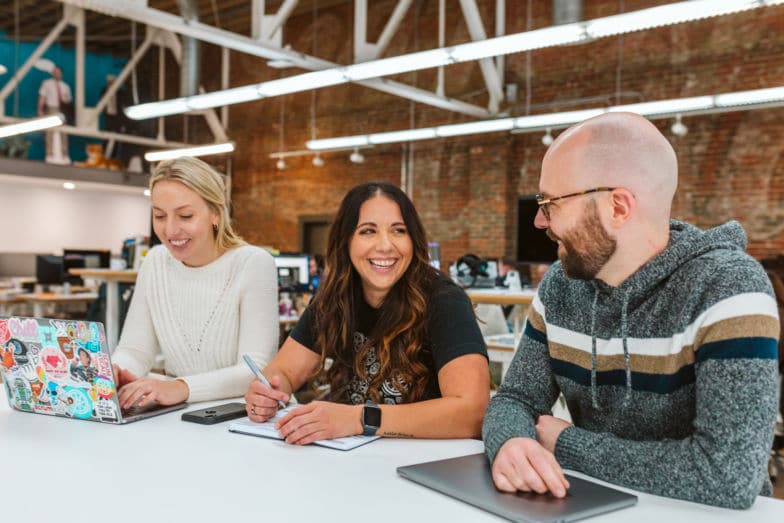 Tre persone sedute una accanto all'altra a un lungo tavolo, tutte sorridenti