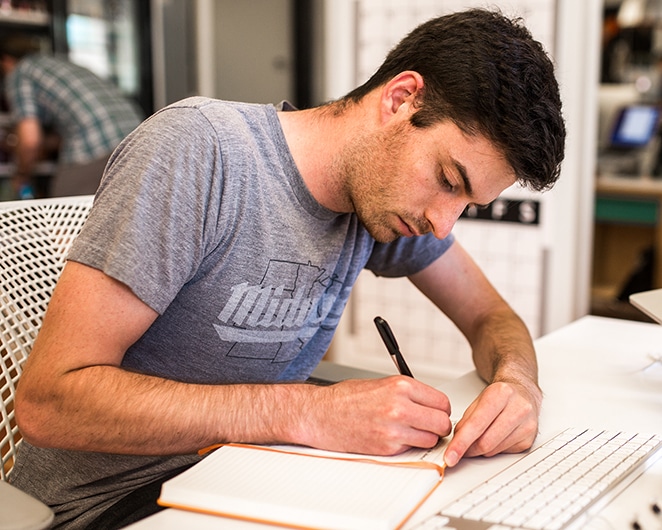 layout per volano ottenere contenuti dai clienti in tempo mantenere i progetti in movimento uomo che indossa una maglietta del Midwest che scrive in un taccuino arancione sulla scrivania bianca